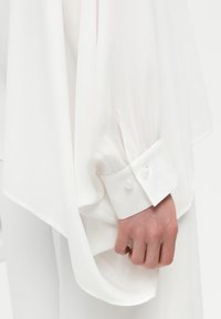 Χέρι με λευκό μπλουζάκι μακρυμάνικο με κουμπωτό μανίκι, χαλαρό ρέον ύφασμα και πιέτες κοντά στον καρπό.