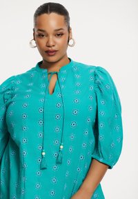 Tegengestelde blouse in turquoise met borduursels, featuring puffmouwen, een keyhole-halslijn met strikjes en bloempatronen in de stof. Zachte textuur.