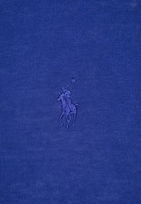 Tissu bleu avec un logo brodé représentant un joueur de polo à cheval, tous deux en fil bleu assorti, centré sur le tissu.