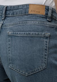 Vue arrière d'une poche de jean en denim bleu avec une étiquette de marque beige indiquant "JDY DENIM" au-dessus de la poche sur une personne portant un haut blanc.