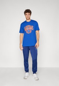 Mėlynas medvilnės marškinėlis su oranžiniu "ADIDAS" spaudiniu, derinamas su tamsiai mėlynomis sportinėmis kelnėmis ir baltais sportiniais batais. Paprastas, laisvo kirpimo dizainas.
