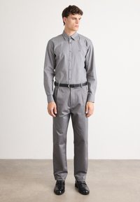 Camicia grigia con colletto e maniche lunghe, abbinata a pantaloni grigi coordinati. Cintura nera e scarpe lucide nere completano l'outfit.