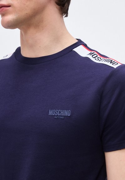 Homme portant un t-shirt Moschino Active bleu marine avec un détail d'épaule marqué blanc et rouge, manches courtes et encolure ronde.