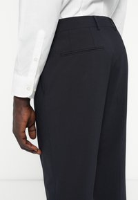 Dunkelblaue Hosen aus glattem Stoff, mit leicht zulaufendem Bein, Gesäßtasche und kombiniert mit einem weißen Langarmhemd.