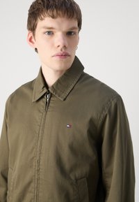 Khaki zip-jakke med spiss krage, to sidelommer og en liten farget logo på brystet. Glatt stofftekstur.