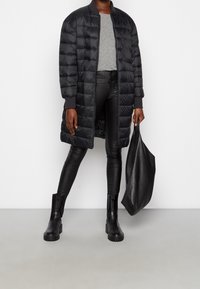 Abrigo acolchado negro con cremallera en la parte frontal y puños de canalé, camisa gris, pantalones de cuero negros y botas negras gruesas, sosteniendo un gran bolso de mano negro.