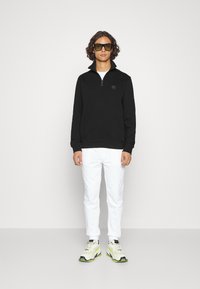 Zwarte rits sweatshirt met een subtiele logo, gecombineerd met witte broeken en chunky sneakers met gele accenten. Staande op een neutrale achtergrond.