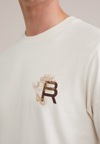 Cream cotton t-shirt featuring a brown stylized "R" graphic and a paisley pattern in gold on the left chest area. Short sleeves, ribbed neckline.
