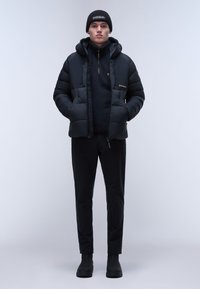 Giacca a vento nera con cappuccio, dotata di una chiusura lampo frontale e tasche laterali, indossata sopra un top in pile nero e pantaloni neri. Berretto nero.