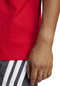 T-shirt rossa di cotone con scollatura a girocollo, abbinata a pantaloni corti neri con strisce diagonali bianche. Mano visibile con un anello decorativo.