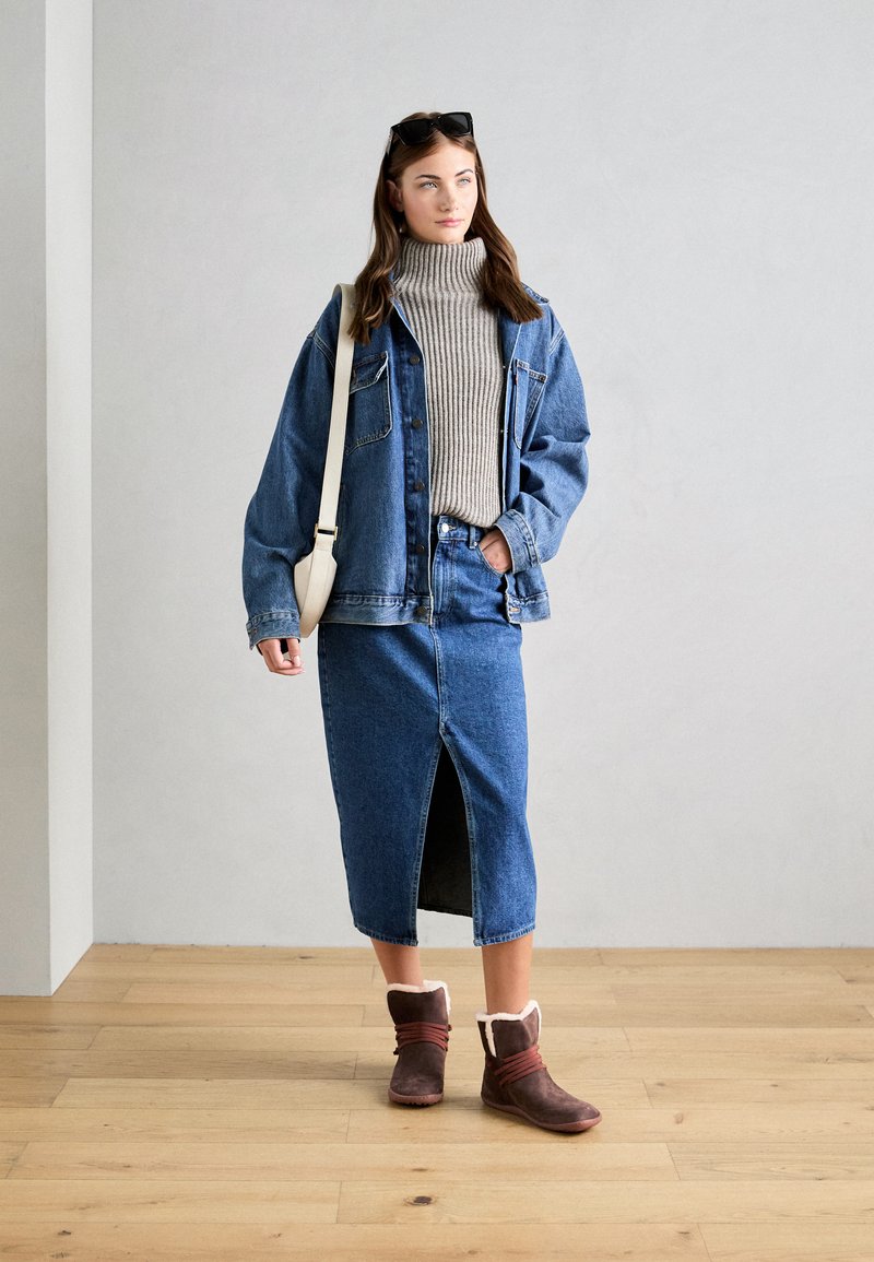 Denimjakke og nederdel med en ribbet grå turtleneck. Brune støvler med pelsfor og røde detaljer. Minimalistisk cremefarvet taske. Trægulv som baggrund.