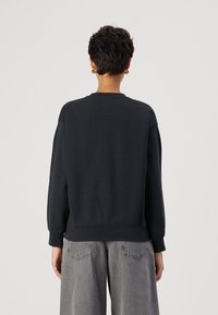 Felpa nera con vestibilità rilassata, scollatura rotonda e polsini a coste. Tessuto liscio, senza motivi visibili, abbinata a pantaloni grigi a gamba larga.