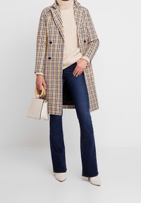 Tvåknäppt rutig kappa i gult, blått och vitt, bärs över en beige stickad tröja, kombineras med utsvängda mörka jeans och krämfärgade klackar.