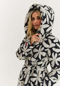 Prošívaný kabát s kapucí, zdobený černo-bílým květinovým vzorem, se zavazováním v pase a texturovanou látkou. Charakteristická přehnaně velká kapuce.