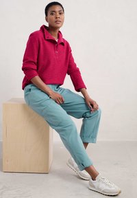 Maglione rosso a trama con colletto alto, abbinato a pantaloni cropped blu chiaro. Le calzature sono sneakers bianche con accenti grigi.