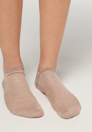 Füße, die beige Unsichtbarkeits-Socken tragen und auf einer schlichten hellen Oberfläche stehen, wobei die Struktur der Socken und die Passform um die Knöchel sichtbar sind.