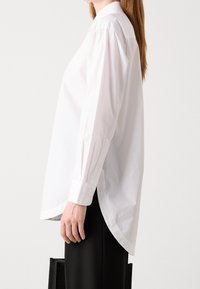 Donna in una camicia bianca larga a maniche lunghe con polsini bottonati e pantaloni neri, che sta in posizione laterale davanti a uno sfondo chiaro e uniforme.