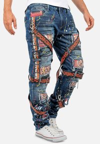 Jean en denim bleu usé avec des éclaboussures de peinture colorées, de multiples déchirures et des sangles décoratives avec du texte. Baskets blanches en dessous.