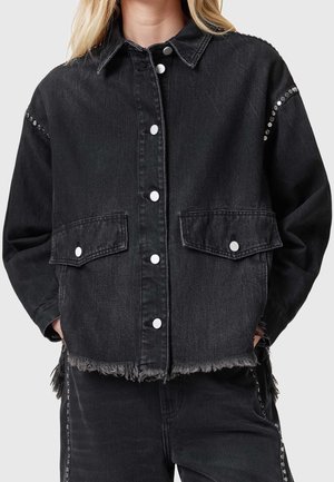 Veste en jean noire avec des boutons-pression argentés, des poches à rabat à l’avant, des détails cloutés sur les manches et un ourlet effiloché portée par une personne les mains dans les poches.