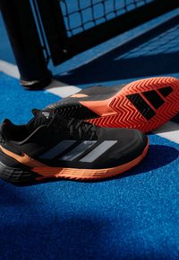 Μαύρα αθλητικά παπούτσια με τρεις ανακλαστικές ασημένιες ρίγες, πορτοκαλί μεσόsole, υφασμάτινη επιφάνεια και μοτίβο από λάστιχο στην σόλα, τοποθετημένα σε μπλε επιφάνεια.