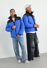 Pufferjakker i lys blå med sorte skuldre, lynlås foran og ribbede manchetter. Modellerne bærer slidte jeans og casual sko.