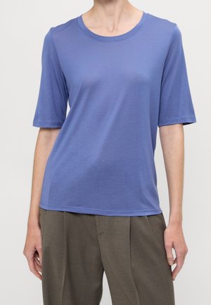 Person wearing a short-sleeve, round-neck periwinkle blue shirt paired with loose-fitting, pleated brown pants against a plain background.