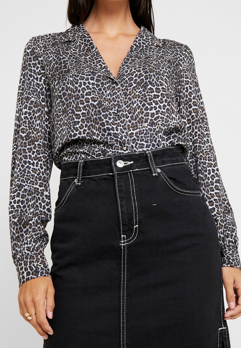 Femme portant un chemisier manches longues à imprimé léopard rentré dans une jupe noire taille haute avec surpiqûres blanches et bouton devant.
