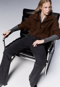 Ruskean mokkanahkajakko, harmaa ribattu villapaita, mustat farkut, istuu mustalla nahkatuolilla. Sormissa renkaita, tyylikäs metallinen tuolin käsivarsi.