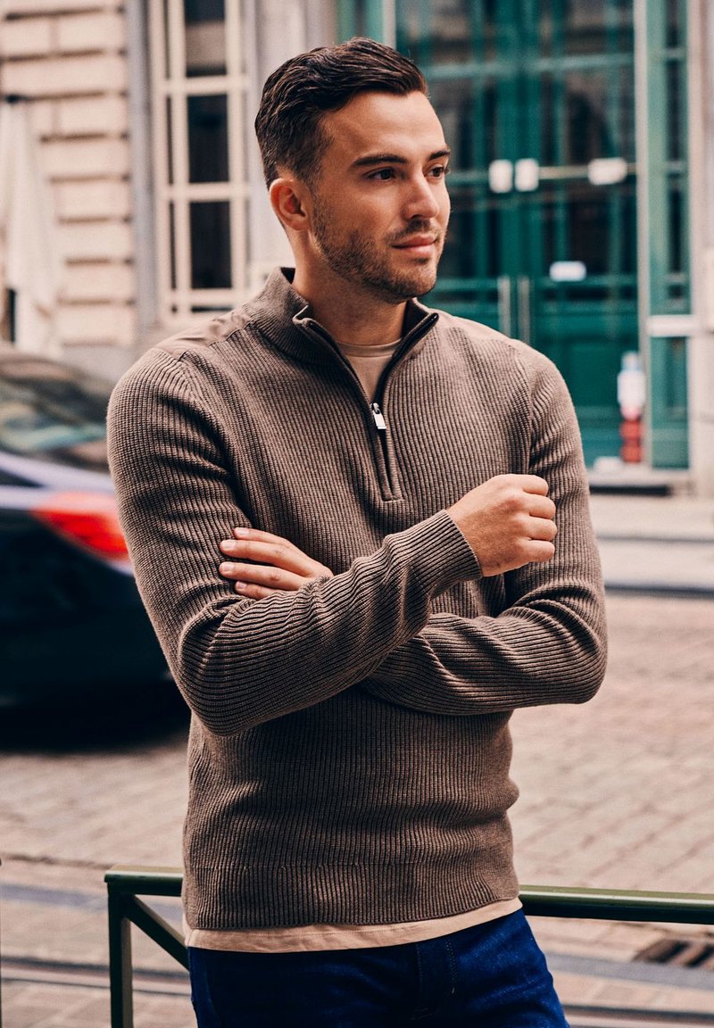 Bruine geribbelde trui met een ritskraag, gedragen over een licht shirt; heeft lange mouwen en een gestructureerde stof. De achtergrond bevat vervaagde auto’s.