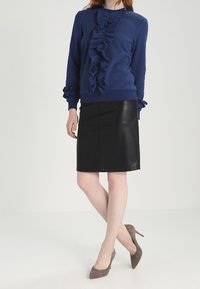 Žena oblečená v námořnicky modré blůze s volánky, černé kožené sukni a šedých semišových lodičkách, stojící s překříženými nohami na bílém pozadí.