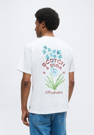 Homme portant un t-shirt blanc avec un motif botanique bleu et vert et coquillages, avec un texte rouge "Scotch & Soda 1985 Amsterdam" dans le dos.