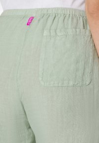 Lichtgroene linnen broek met een elastische tailleband. Bevat een enkele achterzak en een klein paarse merklabel. Zachte, gestructureerde afwerking.