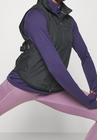 Svart zip-up väst över en lila långärmad tröja, med hög krage, justerbara sidostrapar och en zip-försedd bröstficka.