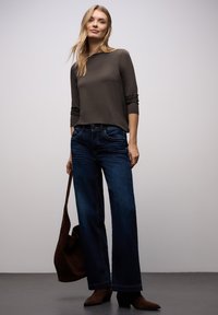 Dunkelbraunes Langarmoberteil kombiniert mit dunkelblauen, weit geschnittenen Denim-Jeans. Das Model trägt eine braune Ledertasche. Der Boden ist grau, der Hintergrund minimalistisch.