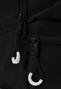 Yksityiskohta mustasta kangaskuosista repusta, jossa on kaksi vetoketjun kahvaa valkoisilla kumikoristeilla, asetettuna tekstuuripohjalle.