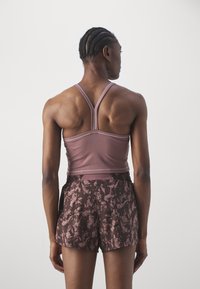 Sportbeha in lichtroze stof met dunne bandjes, gecombineerd met donkerbruine shorts met een textuur van een bloemmotief. Model met de achterkant naar de camera.