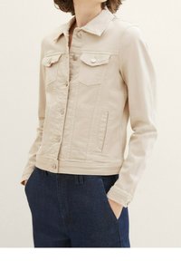 Beige denim jacket with a collar, front button closure, two breast pockets, and side hand pockets. Smooth texture, fitted design.