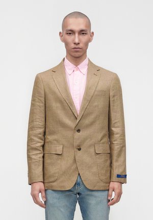 Homme portant un blazer à carreaux beige sur une chemise rose rayée à boutons et un jean bleu clair, debout devant un fond blanc uni.