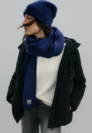 Blue knit beanie, matching blue scarf, and a white ribbed sweater under a black puffer jacket. Gray jeans complete the layered look.