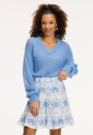 Light blue knitted sweater with a V-neck and loose sleeves paired with a white skirt featuring blue palm tree patterns and embroidered details.
