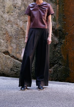 Femme portant un haut en cuir bordeaux à manches courtes, un pantalon large noir et des chaussures noires à bout carré, debout contre un mur en pierre texturé.