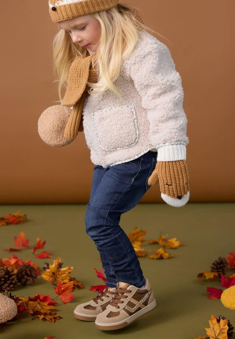 Jeune enfant portant un bonnet en tricot beige, une écharpe et des moufles avec une veste en polaire et un jean, debout parmi les feuilles d’automne et les pommes de pin.