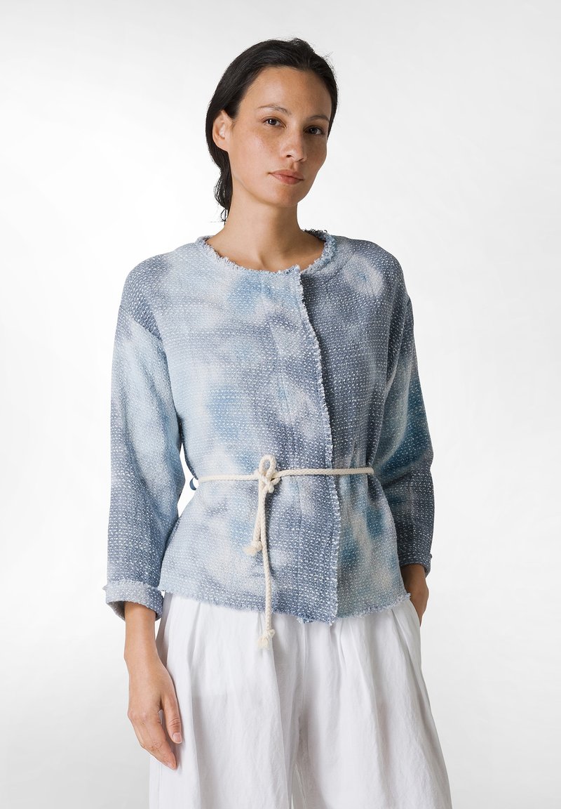 Giacca in tessuto blu chiaro, con texture, dal colletto e dai bordi leggermente sfilacciati, chiusa in vita con un nodo in corda beige, maniche lunghe.