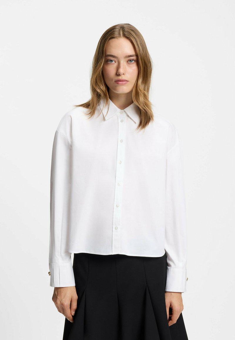 White button-up shirt with a collared neck, dropped shoulders, and long sleeves. Features a cropped design and gold button accents on cuffs.