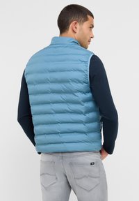 Mann in einer hellblauen gesteppte Weste über einem schwarzen Langarmshirt und hellgrauen Jeans, mit dem Rücken zu einer schlichten Hintergrund.