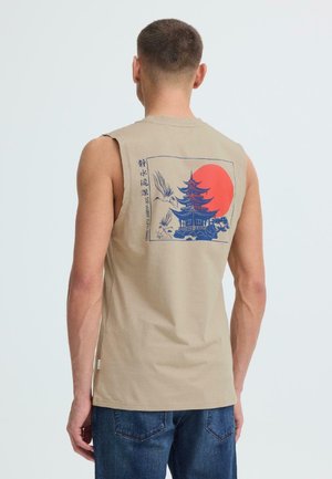 Homme portant une chemise sans manches beige avec une pagode bleue, un soleil rouge, des grues, et le texte « L'eau calme est profonde » au dos, associée à un jean bleu.