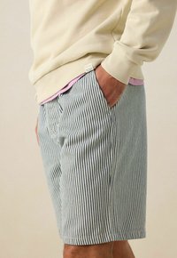 Sweatshirt beige clair sur une chemise rose, associé à un short rayé bleu et blanc avec des poches latérales et une étiquette sur la ceinture.