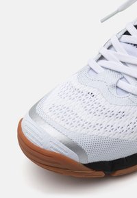 Scarpa sportiva bianca con tomaia in mesh, dettagli argento e suola in gomma marrone. Presenta numerosi fori di ventilazione e lacci bianchi piatti.