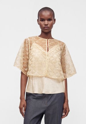 Vrouw met kort haar draagt een doorschijnende gouden kanten top met geschulpte patronen over een crème hemdje en donkergrijze broek.