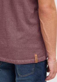 Burgunderfarget bomull t-skjorte med teksturert finish, med en sydd kant og en brun skinn merkehette på siden. Brukt med blå jeans.